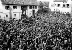 Schwarzweiss Bild, viele Menschen sind auf einem Platz und heben die Hand zum Abstimmen.